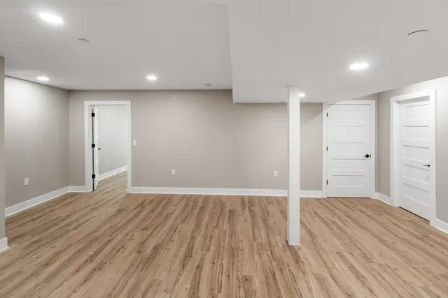 White support column in an open basement with recessed lights, wood-look flooring and paneled doors.
