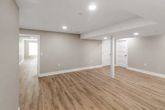 Wide basement view with wood-look flooring, a white support column, paneled doors and a sunlit window.