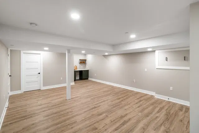 Open basement with wood-look flooring, recessed lights, a high wall window and a beverage nook in the background.