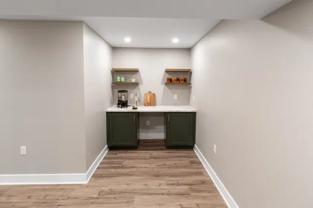 View down the basement beverage alcove toward dark cabinets, a white countertop and open wood shelves.