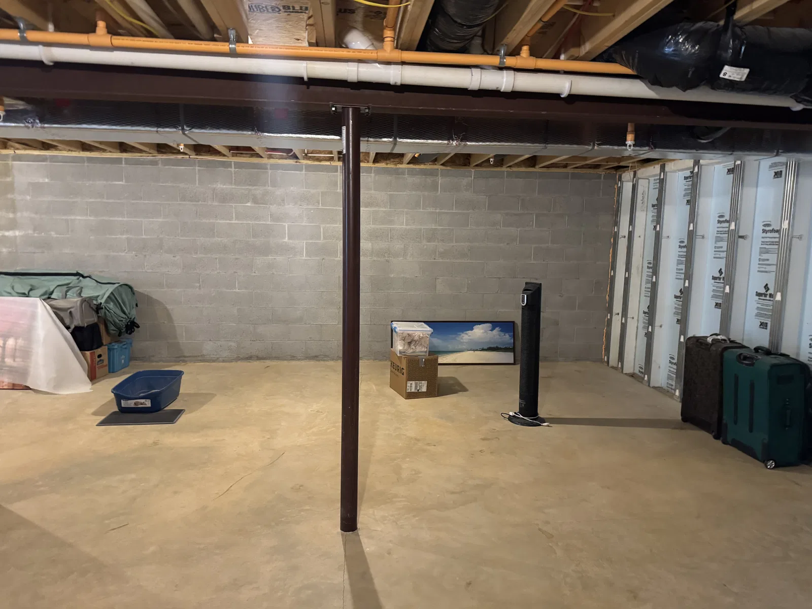 Before finishing: bare block basement wall, concrete floor and steel support post beneath exposed pipes and ductwork.
