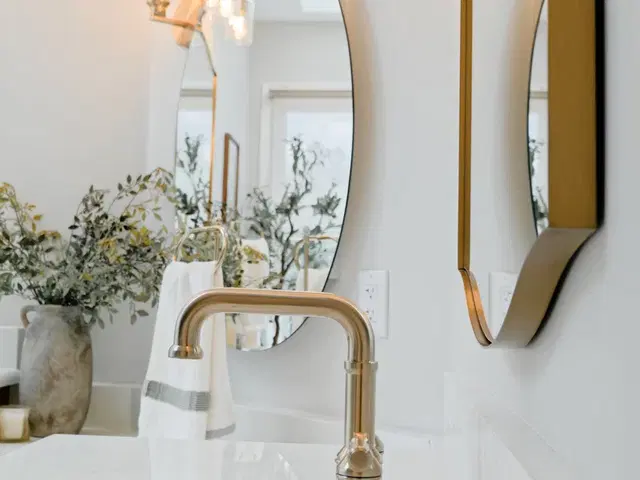 Bathroom sink beneath an arched mirror with gold-tone lighting and a vase of greenery.