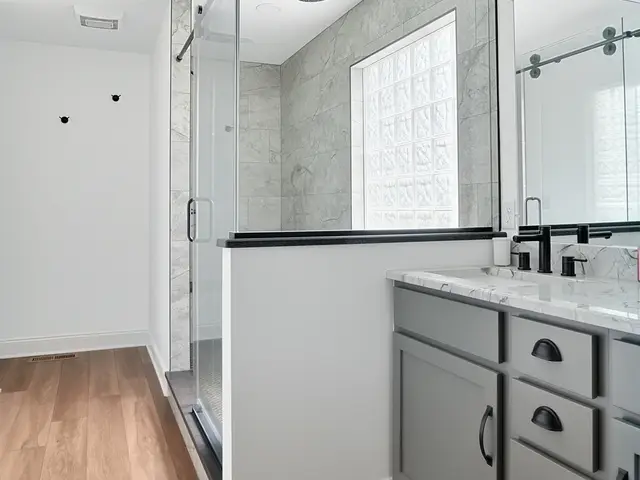 Vertical view of the finished bathroom showing gray vanity cabinets, a glass shower enclosure and wood-look flooring.