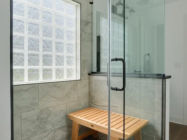Glass shower enclosure with a wood bench, gray tile walls and dark herringbone floor tile.
