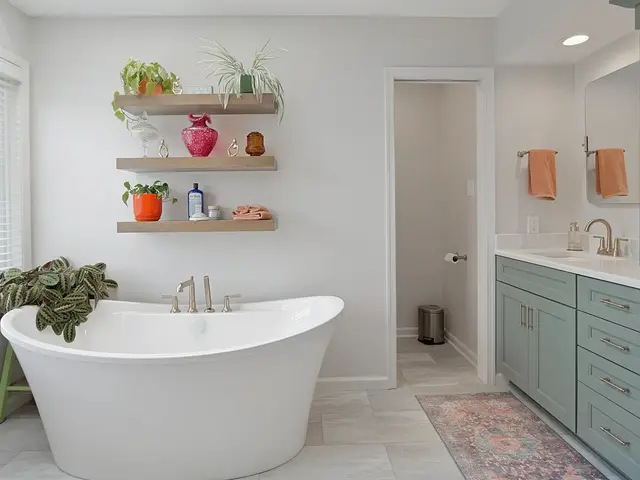 Freestanding white bathtub beside floating wood shelves and a blue-green vanity with gold-tone hardware.