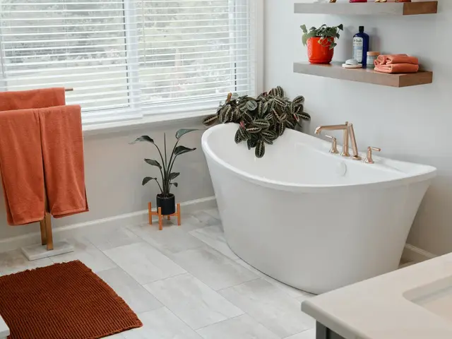 White freestanding tub beneath a window beside floating shelves with plants and folded towels.