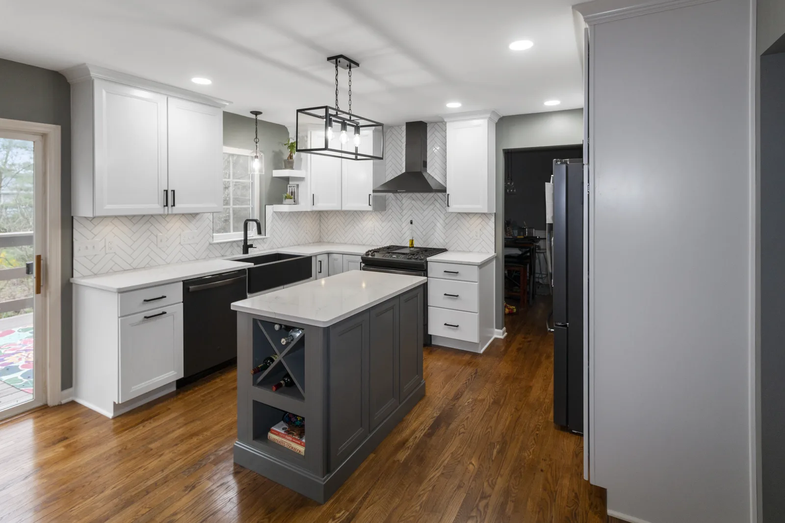 Finished kitchen with white cabinets, a gray island with a wine rack, black apron-front sink, herringbone backsplash and rectangular pendant light.