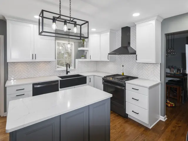 White kitchen cabinetry and a gray island with light veined countertops beside a black sink, gas range and black vent hood.