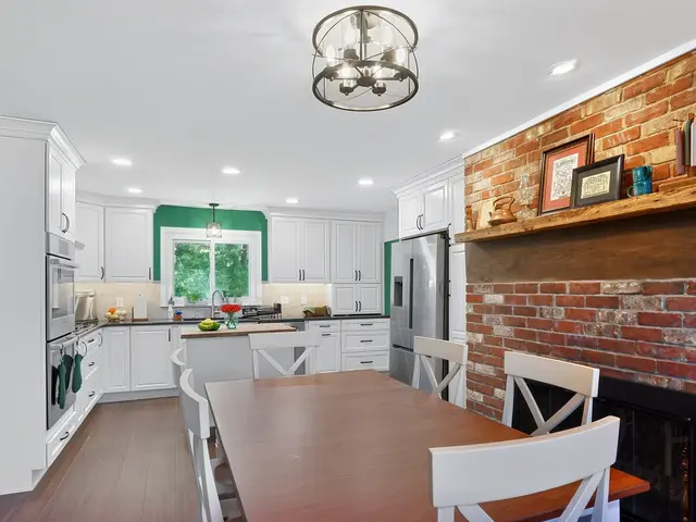 Overview from the dining table toward the white kitchen and wood-topped island, with a brick fireplace and wood mantel along the right wall.