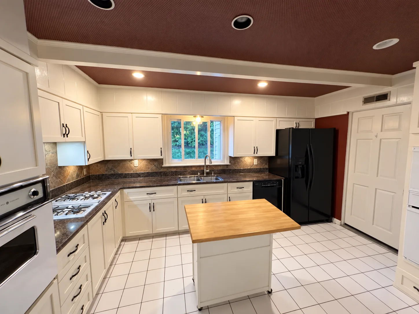 Before remodeling: wide Harrisburg kitchen view showing white cabinetry, dark countertops, a small wood-topped island, black appliances and a brown ceiling with white beams.