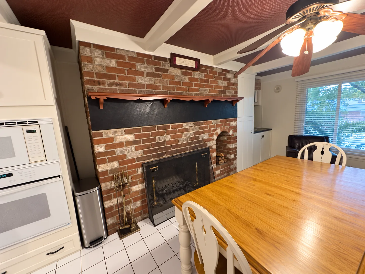 Before remodeling: brick fireplace with a narrow wood mantel and black inset panel beside a white microwave-and-oven cabinet and wood dining table.