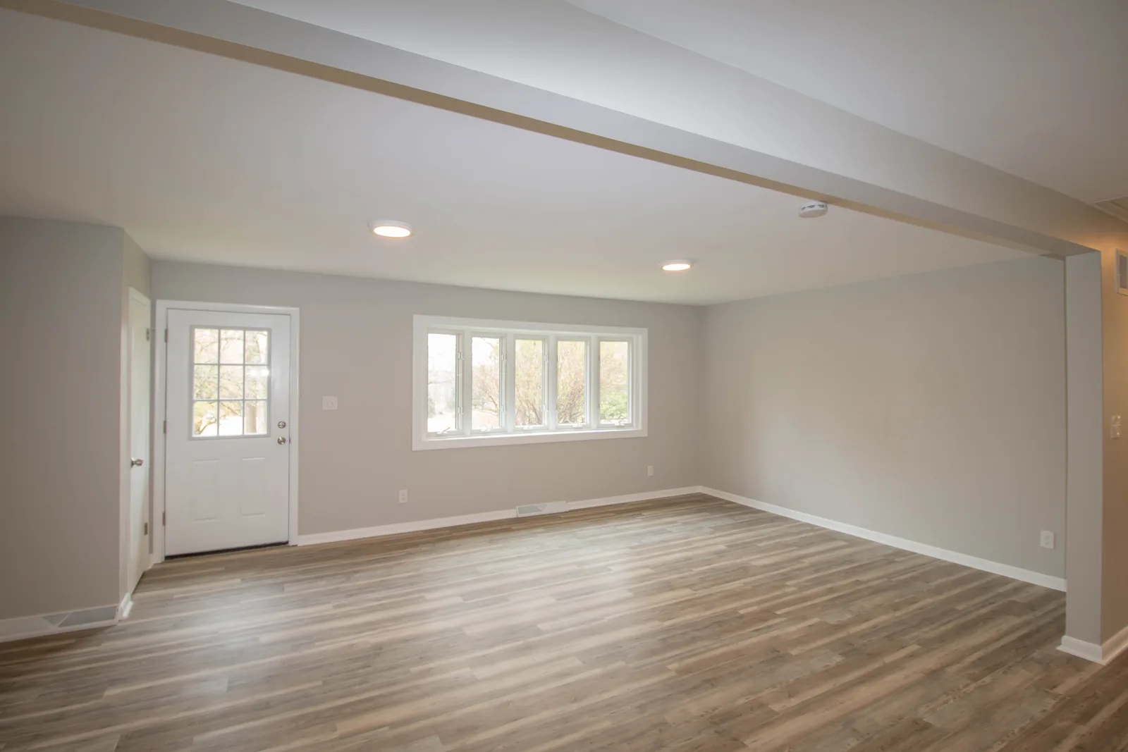 Before remodeling: unfurnished adjoining room with light gray walls, wood-look flooring, a white exterior door and a bank of windows.